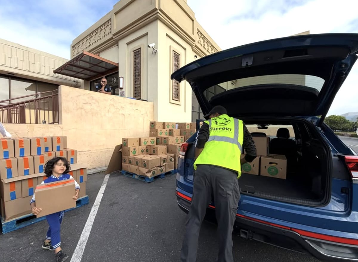 A young boy carries a grocery box while a Support Life Foundation volunteer in a safety vest loads more boxes into a car outside a community center