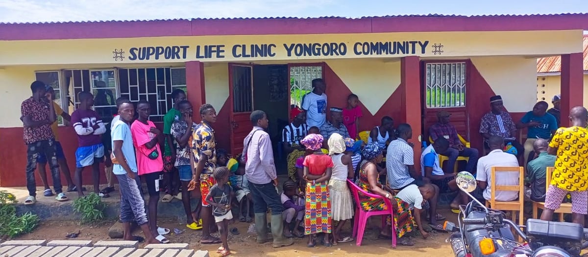 A crowd waiting outside a red and cream building signed Support Life Clinic Yongoro Community, some seated on plastic chairs