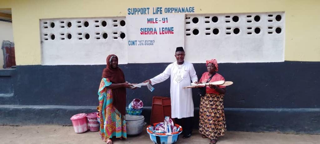 Three adults standing in front of a wall painted Support Life Orphanage, Mile 91, Sierra Leone, handing over cookware, buckets and supplies