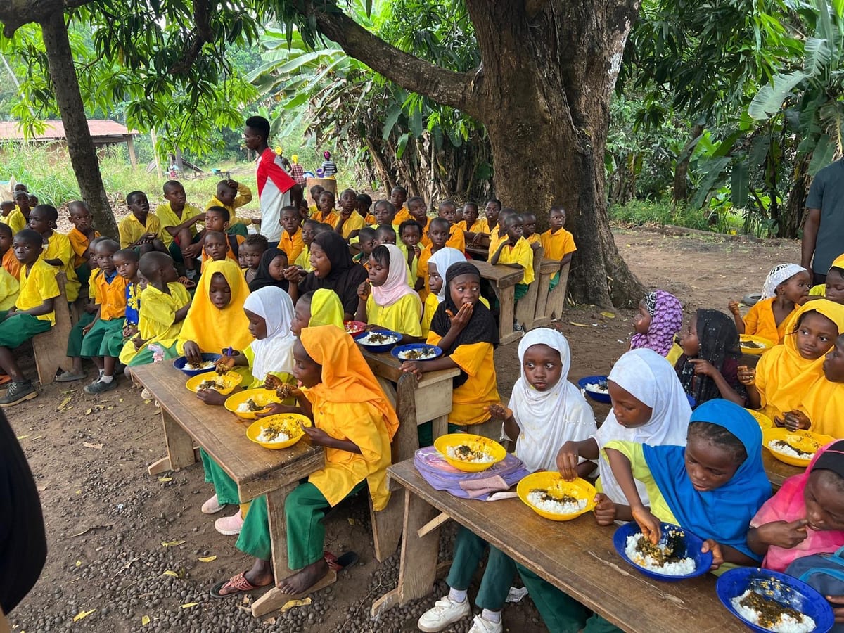 Children in yellow school uniforms seated at benches, eating together from plates at an iftar meal