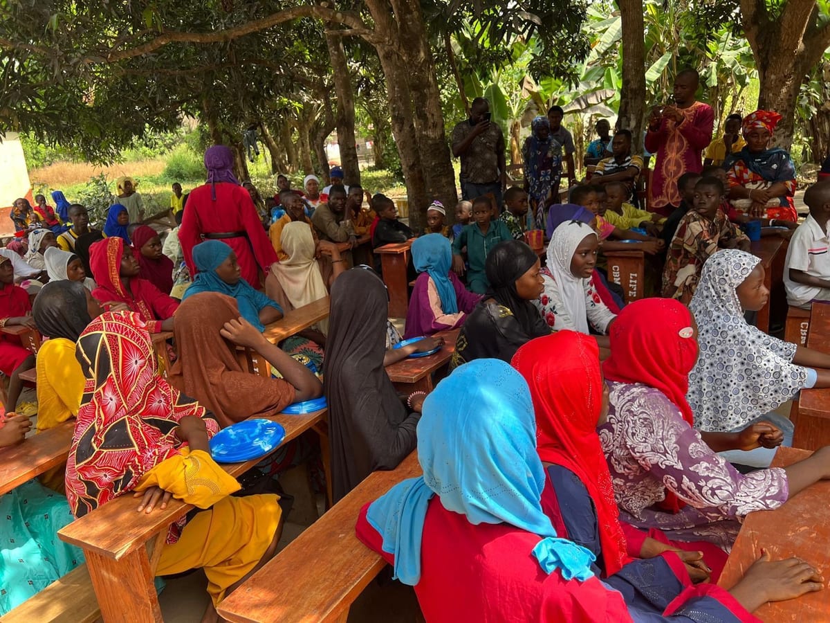 A large gathering of families and children seated at long wooden benches under trees, plates in front of them, waiting to break their fast