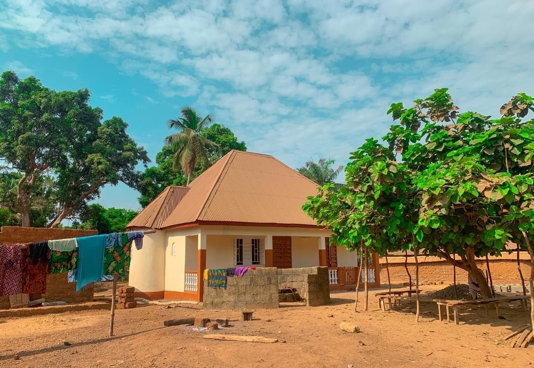 A wide view of the village, low buildings scattered among palm trees on red earth