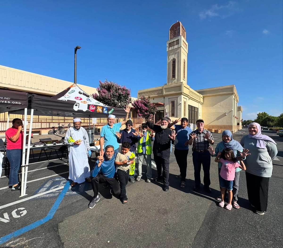 Volunteers and families of all ages, including children, waving together beside the Support Life Foundation tent in a mosque car park on a clear morning